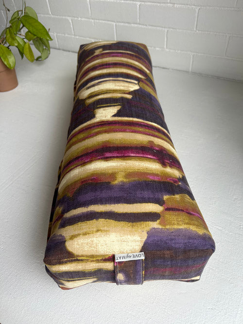 Colorful patterned bolster on a white floor with a plant in the corner.