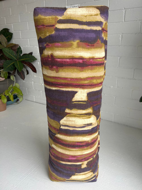 Decorative cylindrical pillow with purple and gold pattern on a white surface.