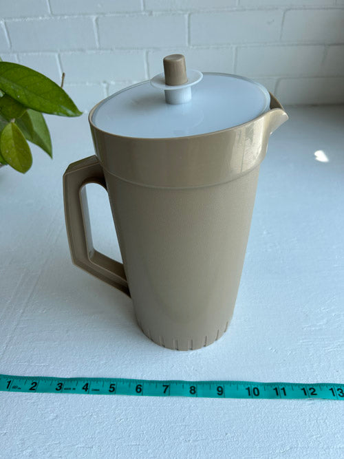 Beige plastic pitcher with a white lid on a white surface, measured by a green tape measure.