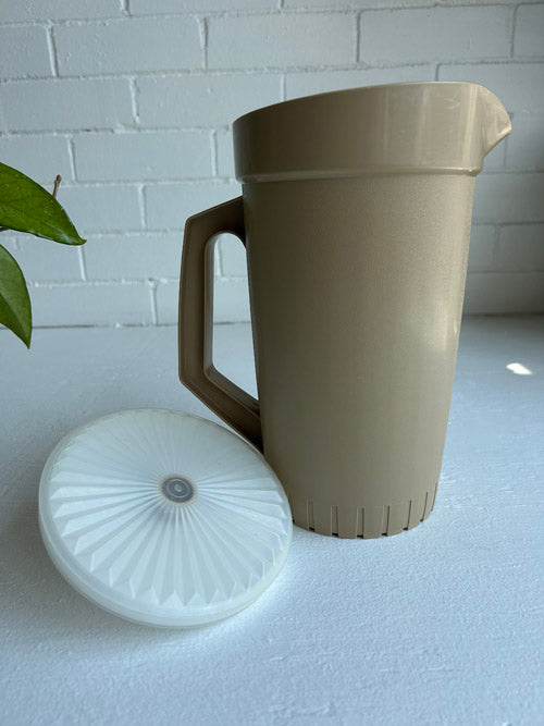 Beige pitcher with a white lid on a light surface with a white brick wall background