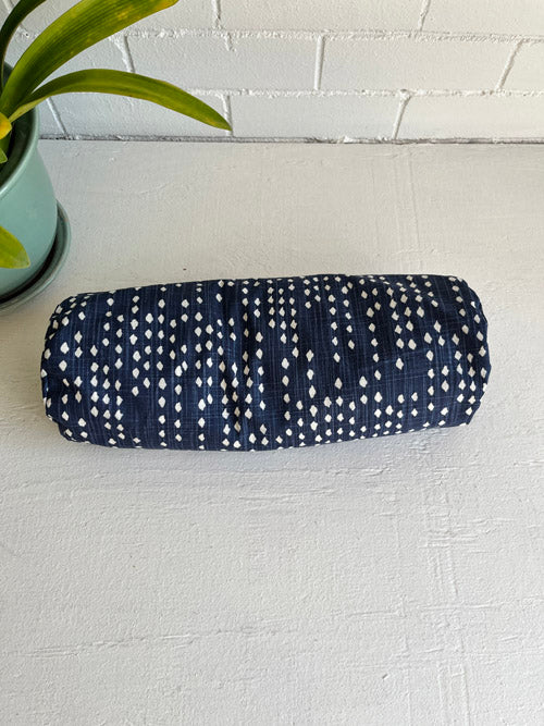 Round yoga bolster in navy blue fabric with white diamond patterns on a white surface, and a plant next to it.