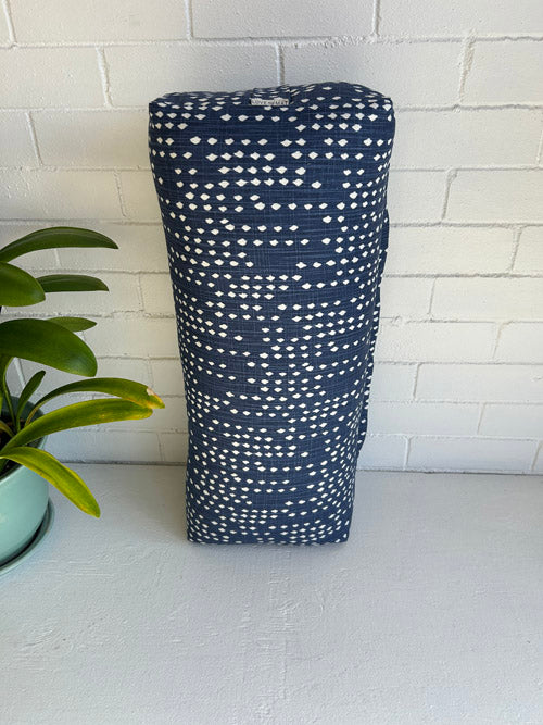Blue and White dotted print Yoga bolster standing on end with a plant next to it and a white brick wall.