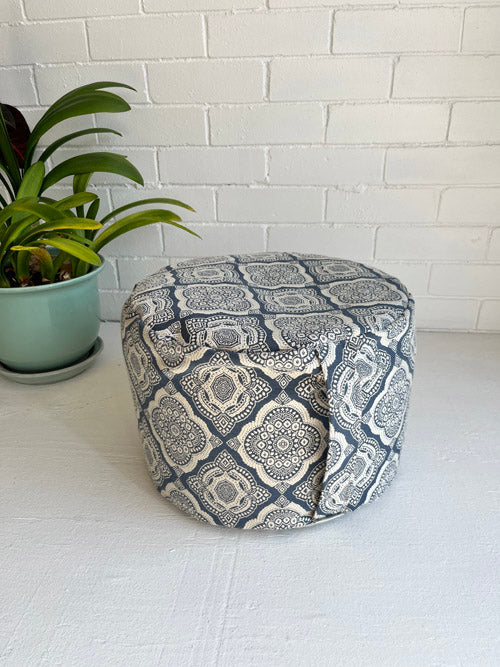 Meditation floor cushion with handle in a geometric pattern on a white surface with a plant in the background