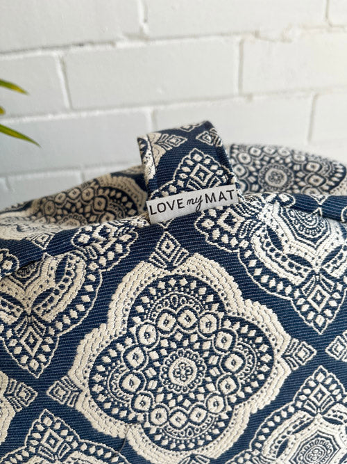 Close up of a navy and white patterned meditation floor pillow with a handle  against a white painted brick background.