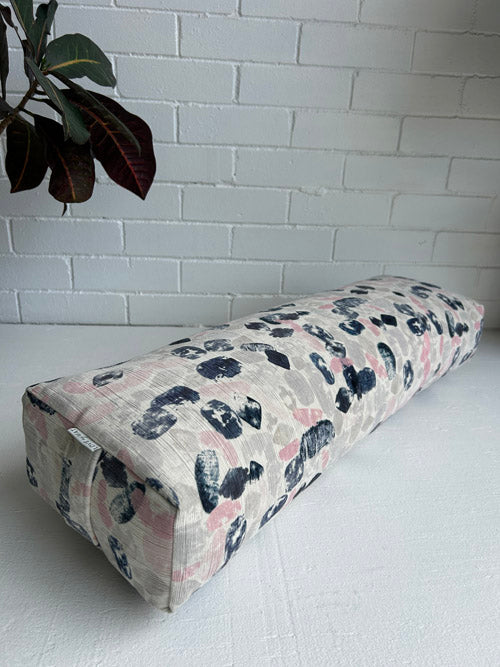 Fabric yoga bolster with black floral pattern on a white surface against a white brick wall.