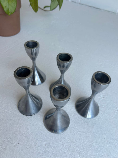 Set of five metallic candle holders on a white surface with a plant in the corner.