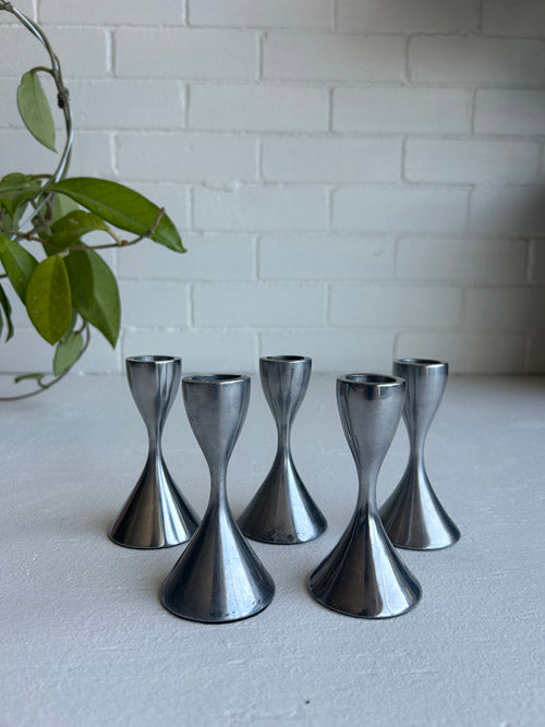 Set of of five metallic candle holders on a light surface with a white brick wall background.
