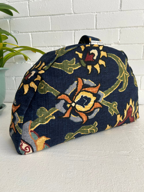 Half moon shaped Meditation pillow sitting up with a handle and a plant nearby against a white brick wall and on a white surface.