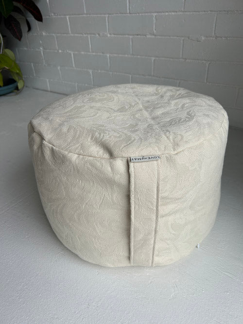 Textured meditation cushion with a visible brand label on a gray floor and brick wall background