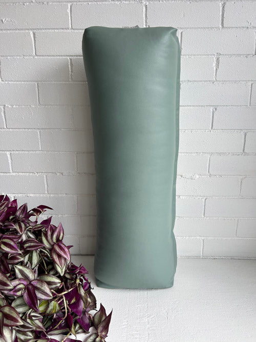 Green long yoga bolster against a white brick wall with a plant on the left.