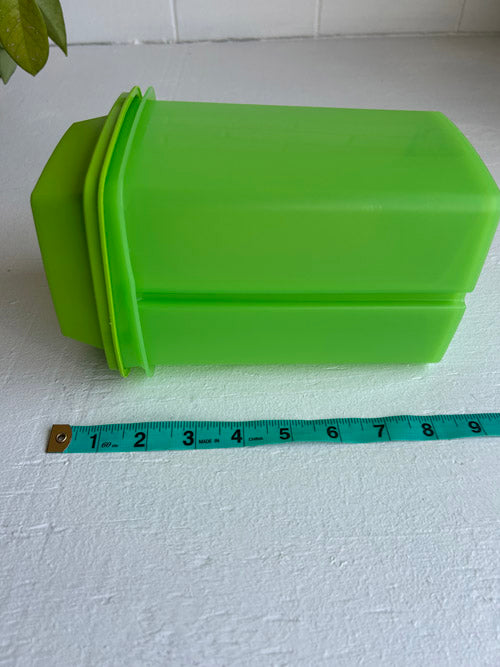 Green plastic container with a green lid on a white surface, measured by a blue tape measure.