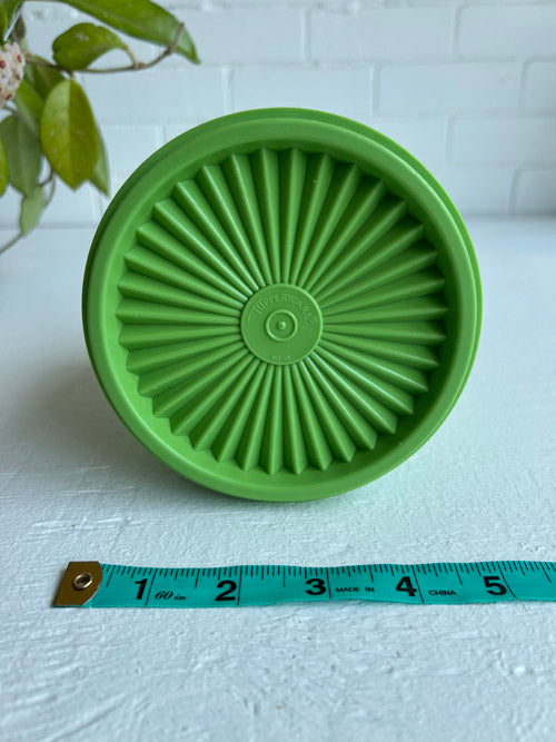 Green silicone lid with sunburst pattern on a white surface, measured against a tape measure.