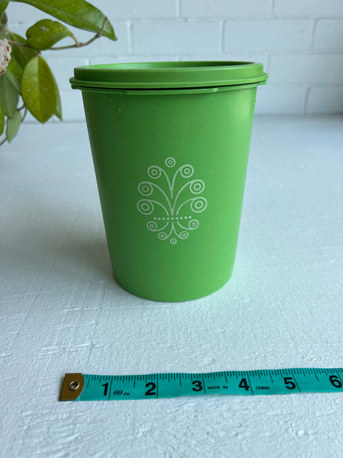 Green canister with a decorative logo on a white surface, accompanied by a measuring tape for scale.