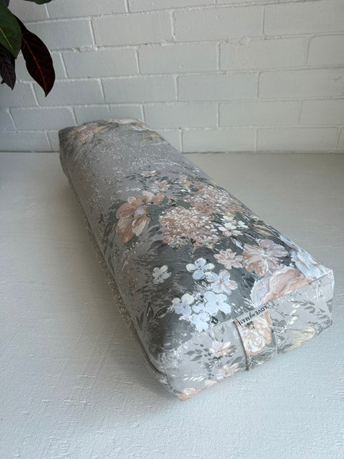 Floral-patterned long sustainable yoga bolster on a white surface with a light gray tiled wall background.