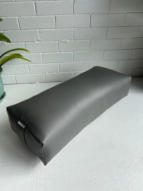Gray traditional yoga bolster on a white surface with a light gray brick wall in the background