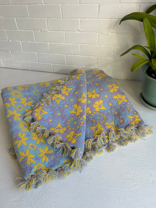 Blue and yellow patterned blanket with tassels on a white surface, next to a plant.