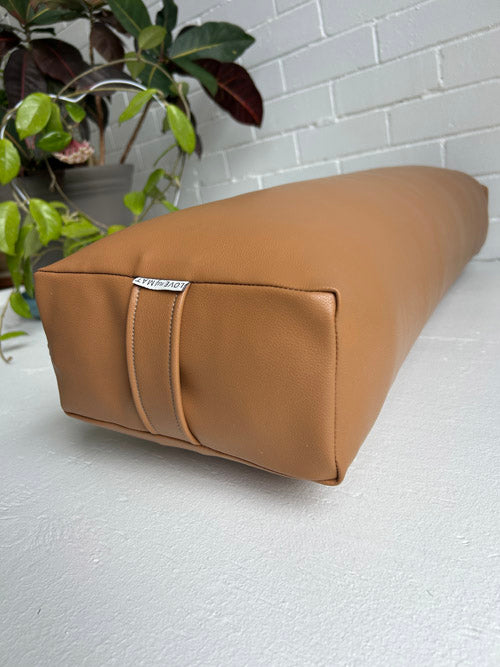 Brown long vinyl yoga bolster on a light gray floor with a white brick wall and plant in the background