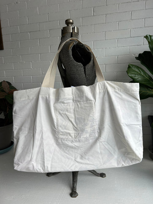 White tote bag with a pocket held by a mannequin against a white brick wall.