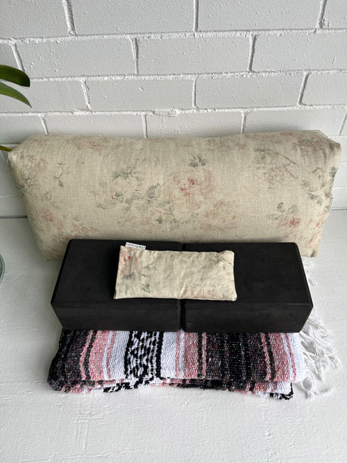 Restorative Yoga Bundle items - bolster, blocks, blanket, and eye pillow on a black bench against a white brick wall.