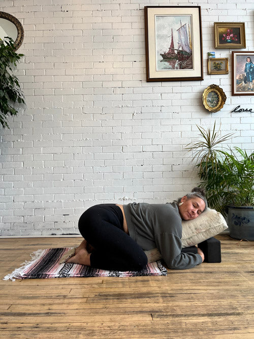 Person lying on a yoga bolster propped up using yoga blocks against a white brick wall with decorative elements.