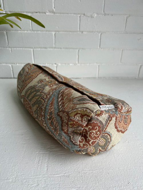Roll-up fabric mini round bolster with paisley pattern on a white surface and light brick wall background