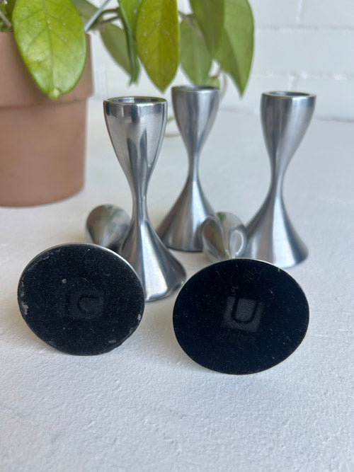 Two black bottoms of candles holders with Umbra logo with 3 others standing in on a white surface, with green leaves in the background.