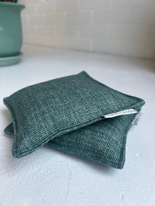Folded green fabric pouch on a white surface with a plant in the background