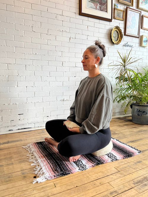 Woman sitting on a yoga mat and sustainable sit cushion in a room with a white brick wall and plants.