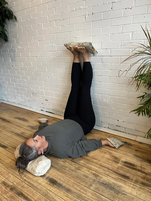 Person lying on a wooden floor with  round bolster under head, knee pillows in hand and sandbag on feet with a white brick wall and plants in the background