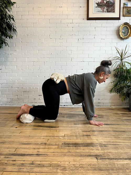 Person practicing yoga with a weighted sandbag on back, mini round bolster under feet and knee pillows under knees on a wooden floor with a white brick wall and plants in the background